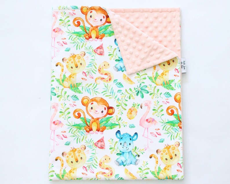 Op de afbeelding: Een wit babydekentje met een jungle dierenprint. Het dekentje heeft apen, luipaarden, een neushoorn, een flamingo en slangen. De achterkant van het dekentje is van zacht, roze minky stof.