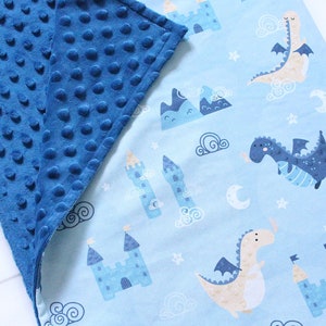 May include: A double-sided blanket featuring a dark blue minky fabric with a raised dot texture on one side and a light blue cotton fabric with a repeating pattern of dragons, castles, mountains, clouds, and moons on the other side with a tag.