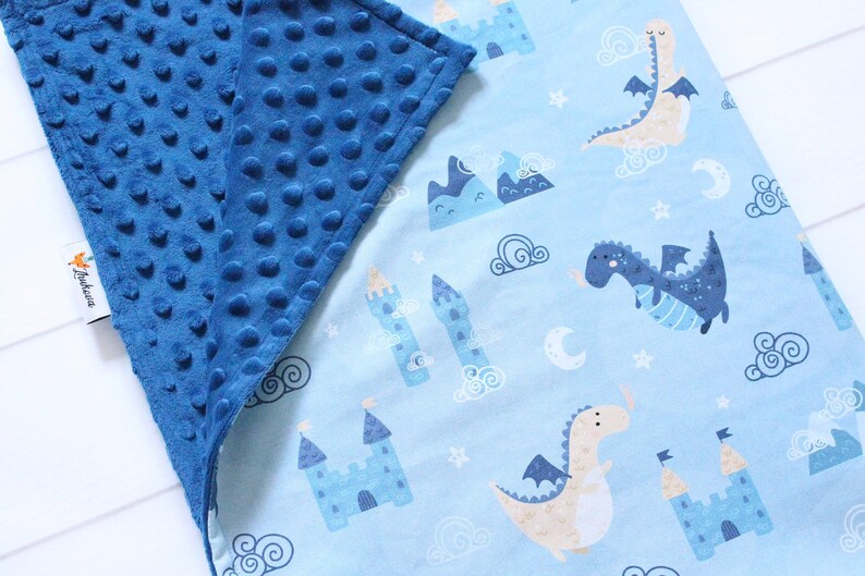 May include: A double-sided blanket featuring a dark blue minky fabric with a raised dot texture on one side and a light blue cotton fabric with a repeating pattern of dragons, castles, mountains, clouds, and moons on the other side with a tag.