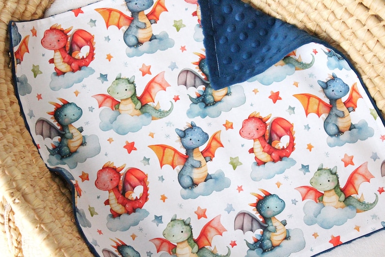 May include: A white baby blanket with a blue minky backing features a repeating pattern of red and blue dragons with stars and clouds.