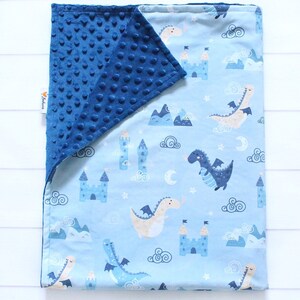 May include: A rectangular baby blanket featuring a light blue side with a repeating pattern of dragons, castles, mountains, clouds, and moons. The reverse side is a solid dark blue minky fabric with a raised dimpled texture. A small white tag is visible.