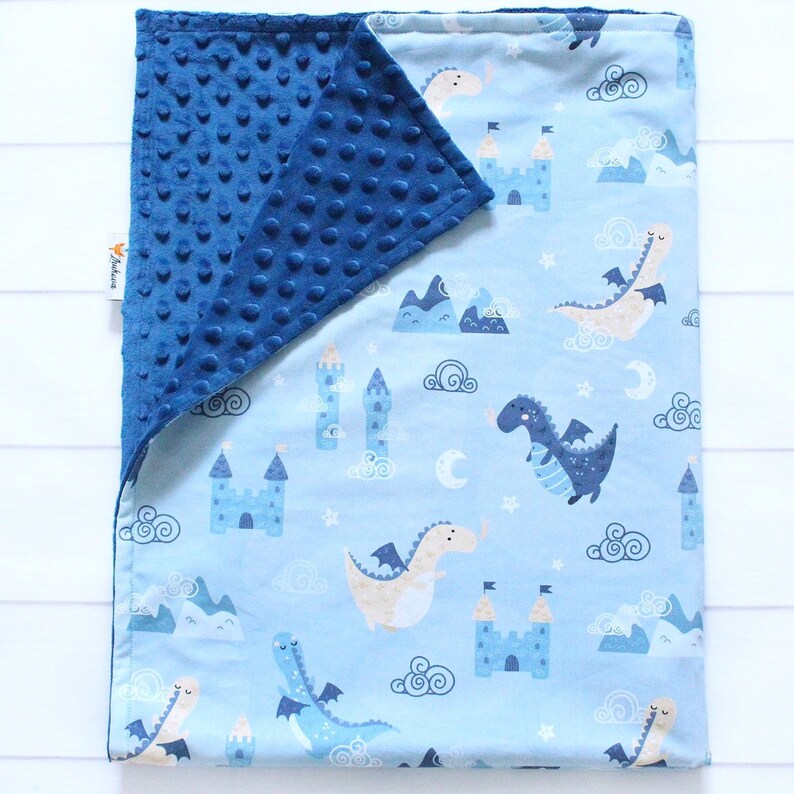 May include: A rectangular baby blanket featuring a light blue side with a repeating pattern of dragons, castles, mountains, clouds, and moons. The reverse side is a solid dark blue minky fabric with a raised dimpled texture. A small white tag is visible.