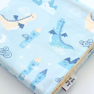 May include: A light blue baby blanket featuring a repeating pattern of cream and blue dragons, blue castles, white crescent moons, stars, and swirling clouds. The blanket has a soft, textured underside and a white fabric tag reading With Love for You.