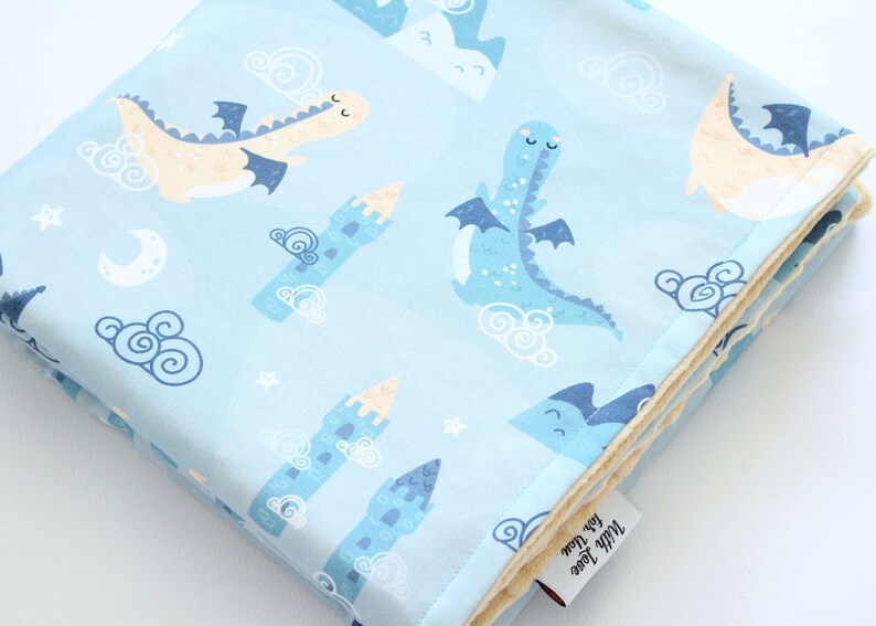 May include: A light blue baby blanket featuring a repeating pattern of cream and blue dragons, blue castles, white crescent moons, stars, and swirling clouds. The blanket has a soft, textured underside and a white fabric tag reading With Love for You.