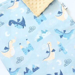 May include: A light blue baby blanket featuring a repeating pattern of dragons, castles, mountains, clouds, stars, and crescent moons. The reverse side is a textured cream-colored fabric with a dimpled surface. A small white tag is visible on the edge.