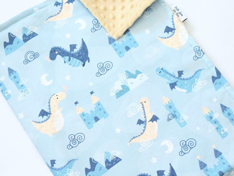 May include: A light blue baby blanket featuring a repeating pattern of dragons, castles, mountains, clouds, stars, and crescent moons. The reverse side is a textured cream-colored fabric with a dimpled surface. A small white tag is visible on the edge.