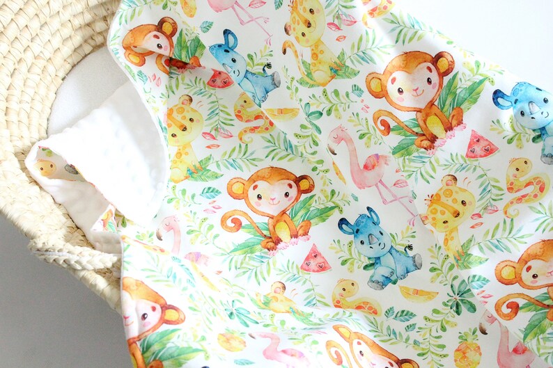 May include: A white fabric baby blanket with a jungle animal print. The print features monkeys, giraffes, rhinos, flamingos, and watermelon slices. The animals are illustrated in a watercolor style.