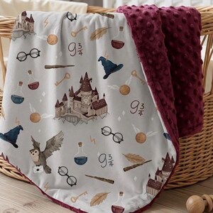 May include: A baby blanket with a light gray background featuring a repeating pattern of a castle, glasses, keys, potion bottles, a wizard hat, an owl, and the number 9 3/4. The blanket has a burgundy, textured backing.
