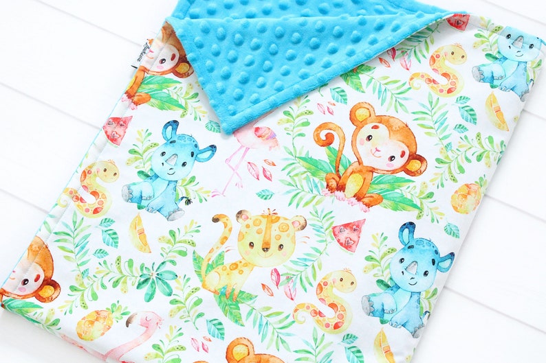 Op de afbeelding: Een witte babydeken met een blauwe minky-rug. De deken heeft een kleurrijke jungledierenprint met apen, neushoorns, slangen en flamingo's.