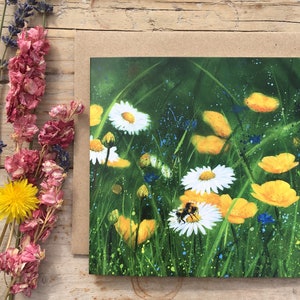 Op de afbeelding: Een vierkante wenskaart met een schilderij van een wilde bloemenweide, met witte madeliefjes, gele boterbloemen en een hommel. De kaart staat tegen een groene achtergrond met blauwe accenten. Gedroogde bloemen zijn op een houten oppervlak geplaatst.
