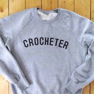 Puede incluir: Una sudadera gris con una impresión negra que dice "CROCHETER" en un arco curvo.