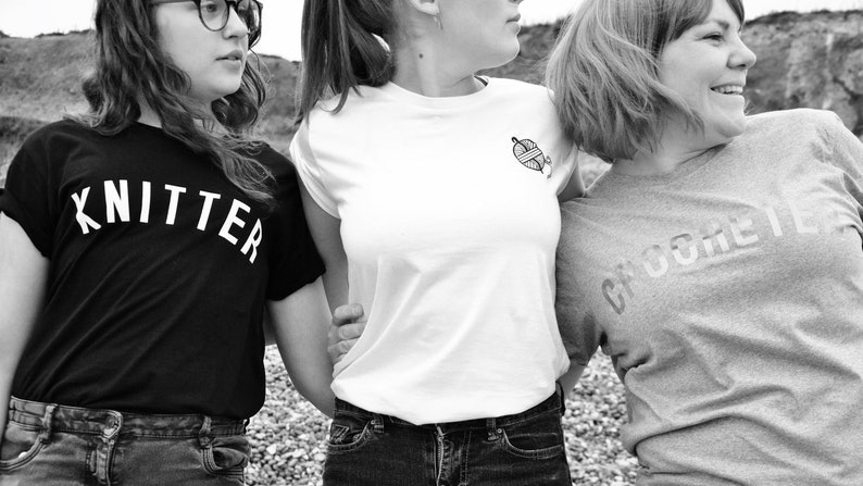 Puede incluir: Tres amigas con camisetas negras, blancas y grises con las palabras "Knitter", "Crocheter" y un gr&aacute;fico de ovillo de lana.