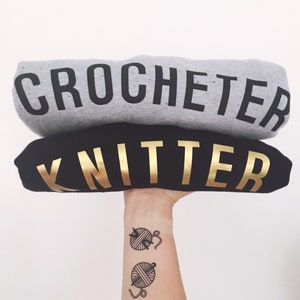 Puede incluir: Dos sudaderas grises y negras apiladas una encima de la otra. La sudadera de arriba dice "Crocheter" en letras negras. La sudadera de abajo dice "Knitter" en letras doradas. Un brazo de una persona con dos tatuajes de ovillos de lana y agujas est&aacute; sosteniendo las sudaderas.