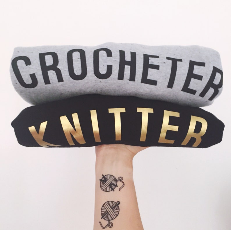 Puede incluir: Dos sudaderas grises y negras apiladas una encima de la otra. La sudadera de arriba dice "Crocheter" en letras negras. La sudadera de abajo dice "Knitter" en letras doradas. Un brazo de una persona con dos tatuajes de ovillos de lana y agujas est&aacute; sosteniendo las sudaderas.