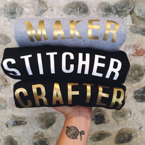 Puede incluir: Tres camisetas negras apiladas una encima de la otra. La camiseta de arriba tiene la palabra "MAKER" en letras doradas. La camiseta del medio tiene la palabra "STITCHER" en letras blancas. La camiseta de abajo tiene la palabra "CRAFTER" en letras doradas.