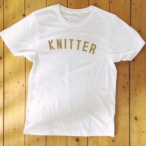 Puede incluir: Camiseta blanca con un estampado dorado de "KNITTER" en la parte delantera.