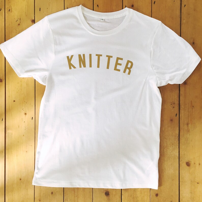 Puede incluir: Camiseta blanca con un estampado dorado de "KNITTER" en la parte delantera.