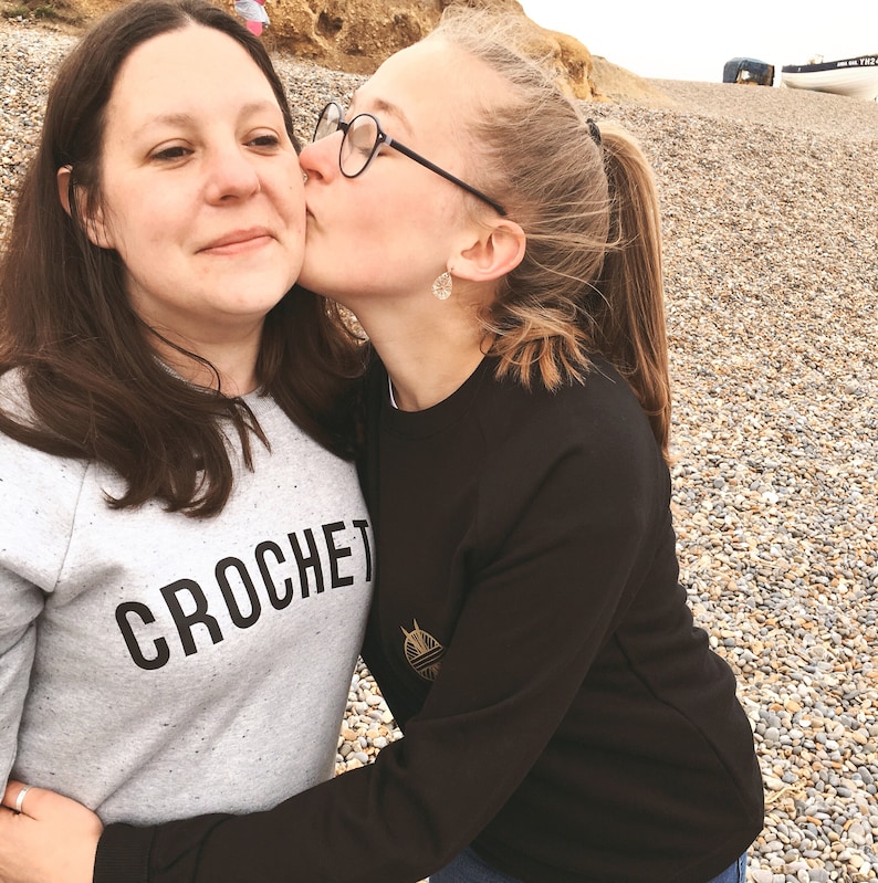 Puede incluir: Dos personas, una madre y su hija, se abrazan en una playa. La hija est&aacute; besando a la madre en la mejilla. La madre lleva una sudadera gris con la palabra "CROCHET" impresa. La hija lleva una sudadera negra con un dise&ntilde;o de ovillo de lana dorado.