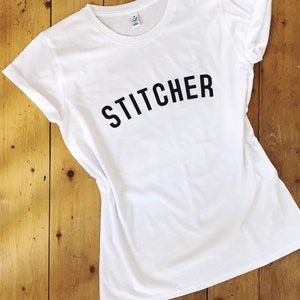 May include: White t-shirt with the word "STITCHER" printed in black on the front.