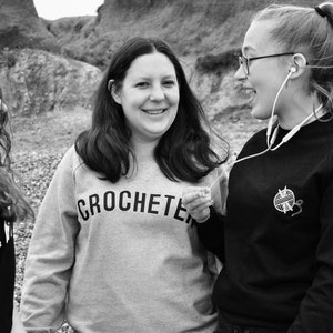 Puede incluir: Tres personas est&aacute;n de pie afuera, dos mujeres sonr&iacute;en y hablan, y una tercera mujer mira hacia otro lado. La mujer del medio lleva una sudadera gris con la palabra "CROCHETER" impresa en ella. La mujer de la derecha lleva una sudadera negra con un peque&ntilde;o dise&ntilde;o blanco de un ovillo de lana y un ganchillo.