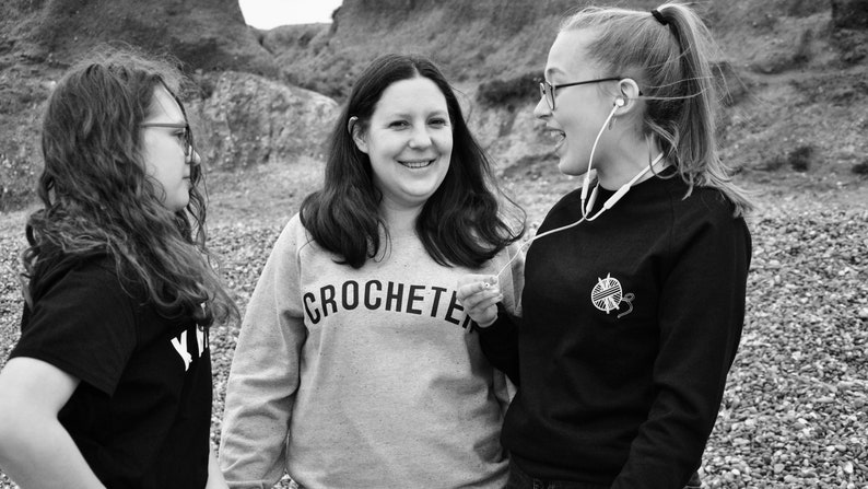 Puede incluir: Tres personas est&aacute;n de pie afuera, dos mujeres sonr&iacute;en y hablan, y una tercera mujer mira hacia otro lado. La mujer del medio lleva una sudadera gris con la palabra "CROCHETER" impresa en ella. La mujer de la derecha lleva una sudadera negra con un peque&ntilde;o dise&ntilde;o blanco de un ovillo de lana y un ganchillo.