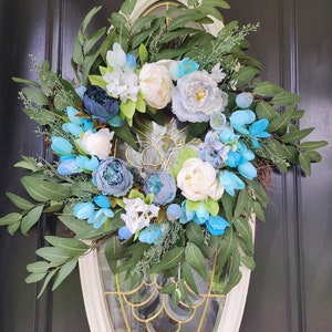 May include: A blue and white floral wreath with green leaves. The wreath is hanging on a black door with a white oval frame.