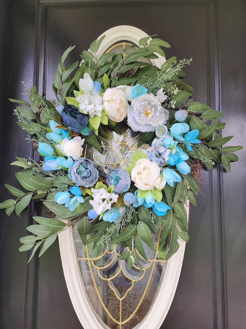 May include: A blue and white floral wreath with green leaves. The wreath is hanging on a black door with a white oval frame.