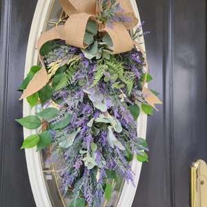 Peut inclure: Une composition florale en forme de goutte d'eau avec de la lavande, du feuillage vert et un n&oelig;ud en toile de jute. L'arrangement est pr&eacute;sent&eacute; sur une porte avec une fen&ecirc;tre ovale. La lavande et la verdure tombent en cascade.