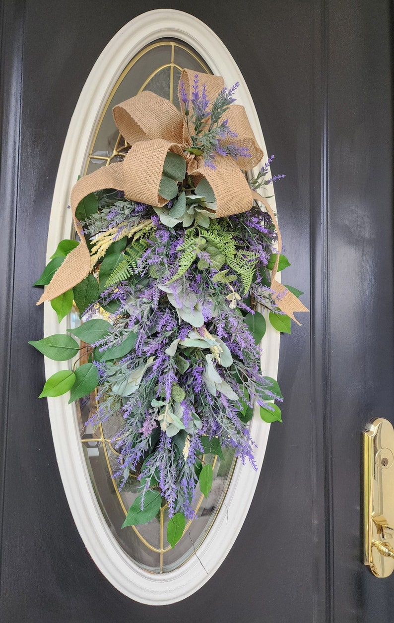 Peut inclure: Une composition florale en forme de goutte d'eau avec de la lavande, du feuillage vert et un n&oelig;ud en toile de jute. L'arrangement est pr&eacute;sent&eacute; sur une porte avec une fen&ecirc;tre ovale. La lavande et la verdure tombent en cascade.