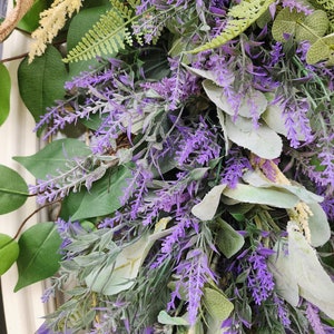 Peut inclure: Gros plan sur une couronne d&eacute;corative avec de la lavande violette vibrante, des feuilles vertes et des accents cr&egrave;me. La couronne est orn&eacute;e de diff&eacute;rents types de feuillage, cr&eacute;ant une apparence naturelle et textur&eacute;e. Un ruban de jute ajoute une touche rustique.