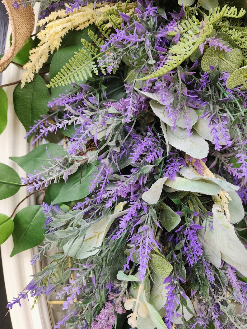 Peut inclure: Gros plan sur une couronne d&eacute;corative avec de la lavande violette vibrante, des feuilles vertes et des accents cr&egrave;me. La couronne est orn&eacute;e de diff&eacute;rents types de feuillage, cr&eacute;ant une apparence naturelle et textur&eacute;e. Un ruban de jute ajoute une touche rustique.