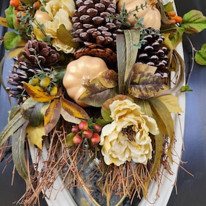 Op de afbeelding: Een herfst-deurkrans met kunstbloemen, dennenappels en pompoenen. De krans is versierd met bruine takken en rode bessen.