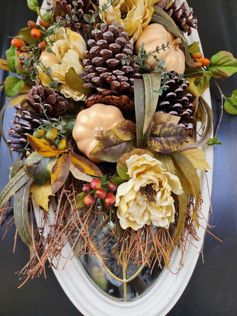 Op de afbeelding: Een herfst-deurkrans met kunstbloemen, dennenappels en pompoenen. De krans is versierd met bruine takken en rode bessen.