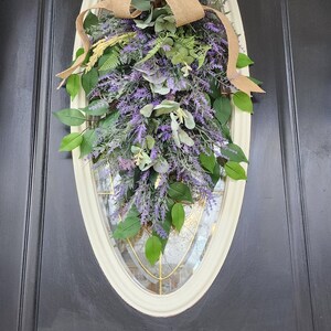 Peut inclure: D&eacute;coration de porte ovale avec un m&eacute;lange de lavande artificielle, de feuilles vertes et d'eucalyptus. Un n&oelig;ud en toile de jute est plac&eacute; en haut. L'ensemble est pos&eacute; sur une porte noire avec un cadre cr&egrave;me.
