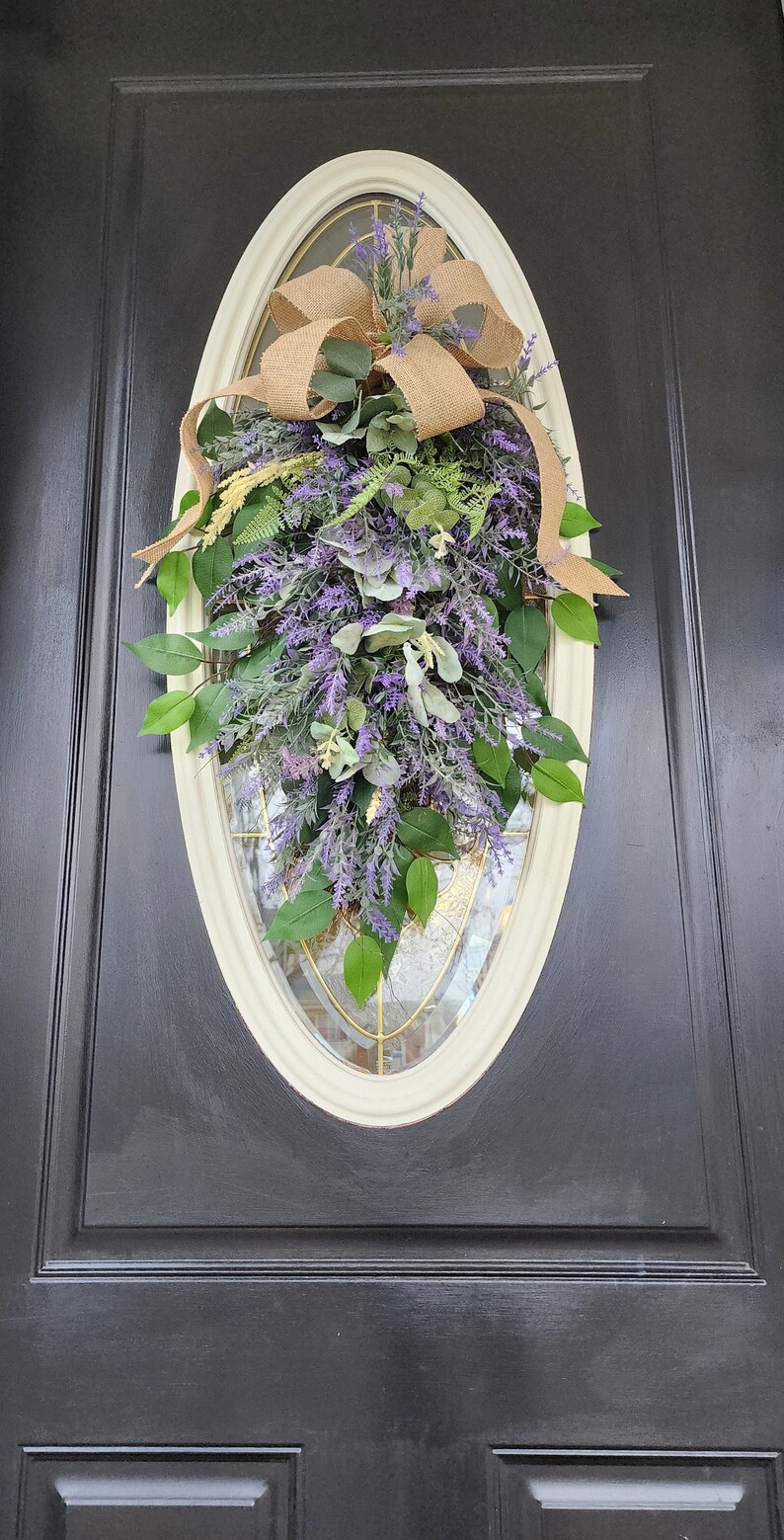 Peut inclure: D&eacute;coration de porte ovale avec un m&eacute;lange de lavande artificielle, de feuilles vertes et d'eucalyptus. Un n&oelig;ud en toile de jute est plac&eacute; en haut. L'ensemble est pos&eacute; sur une porte noire avec un cadre cr&egrave;me.
