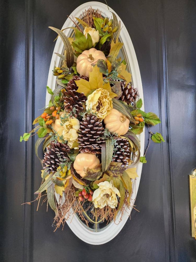 Op de afbeelding: Een herfst-deurkrans met nep-pompoenen, dennenappels en bloemen in tinten goud, bruin en wit. De krans is ovaalvormig en hangt aan een zwarte deur.