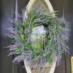 May include: A lavender wreath with purple flowers and green leaves hangs on a black door with a white oval frame.