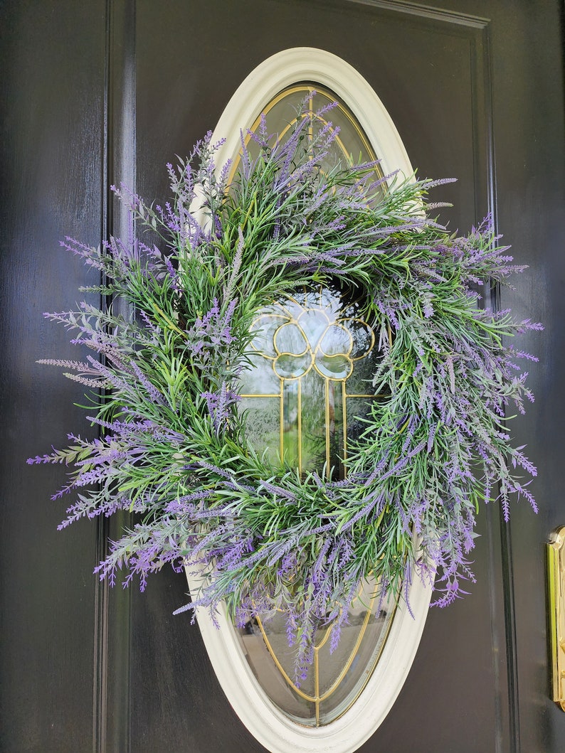 May include: A lavender wreath with purple flowers and green leaves hangs on a black door with a white oval frame.