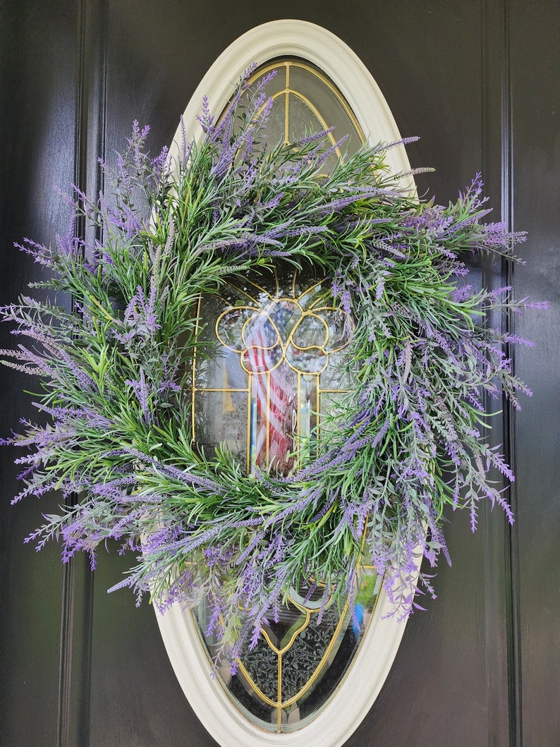 May include: A lavender wreath with green leaves hangs on a black door with a white oval frame and a stained glass window behind it.