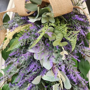 Peut inclure: Une composition florale d&eacute;corative avec de la lavande, du feuillage vert et un n&oelig;ud en toile de jute. L'arrangement comprend diff&eacute;rents types de verdure et des brins de lavande violets, cr&eacute;ant un aspect naturel et textur&eacute;. Le n&oelig;ud en toile de jute ajoute une touche rustique.