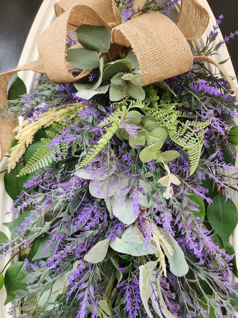 Peut inclure: Une composition florale d&eacute;corative avec de la lavande, du feuillage vert et un n&oelig;ud en toile de jute. L'arrangement comprend diff&eacute;rents types de verdure et des brins de lavande violets, cr&eacute;ant un aspect naturel et textur&eacute;. Le n&oelig;ud en toile de jute ajoute une touche rustique.