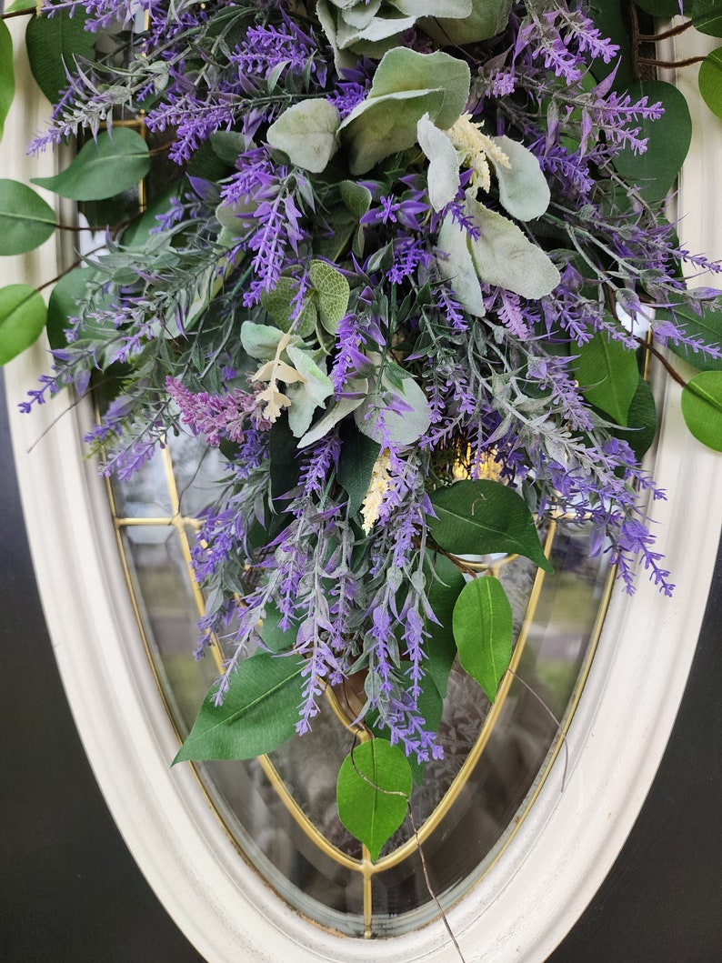Peut inclure: Gros plan sur une composition florale avec des brins de lavande violets, des feuilles vertes et un feuillage gris-vert. L'arrangement est plac&eacute; devant une porte vitr&eacute;e &agrave; cadre blanc. La lavande et la verdure cr&eacute;ent un affichage naturel et accueillant.