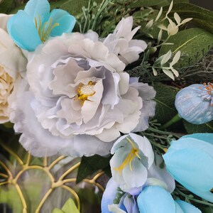 May include: A wreath featuring blue and white flowers, green leaves, and a light blue flower bud. The wreath is crafted from artificial flowers and greenery.