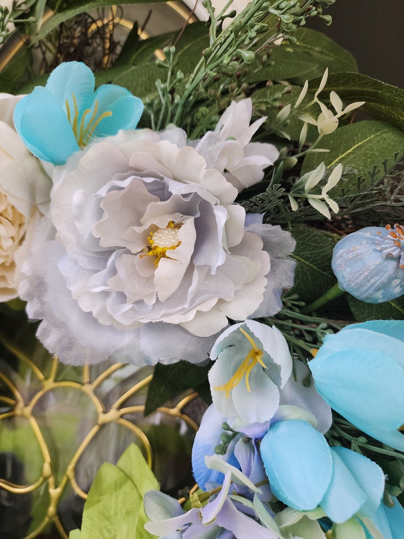 May include: A wreath featuring blue and white flowers, green leaves, and a light blue flower bud. The wreath is crafted from artificial flowers and greenery.
