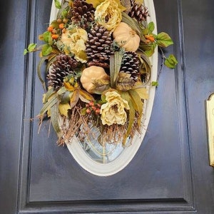 Op de afbeelding: Een herfst-deurkrans met nep-pompoenen, dennenappels en bloemen in tinten bruin, goud en wit. De krans wordt getoond op een zwarte deur.