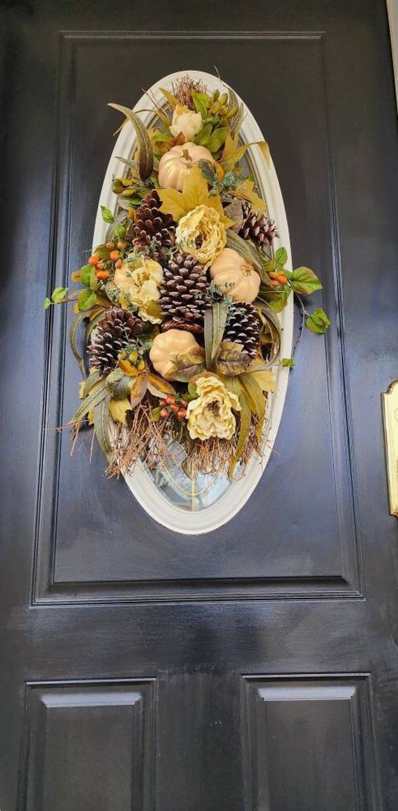 Op de afbeelding: Een herfst-deurkrans met nep-pompoenen, dennenappels en bloemen in tinten bruin, goud en wit. De krans wordt getoond op een zwarte deur.