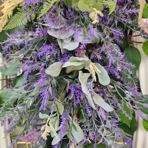 Peut inclure: Gros plan sur une couronne d&eacute;corative avec de la lavande violette, des feuilles vertes et un feuillage cr&egrave;me. La couronne est dens&eacute;ment garnie de diverses textures et nuances de vert et de violet, cr&eacute;ant une esth&eacute;tique naturelle et accueillante.
