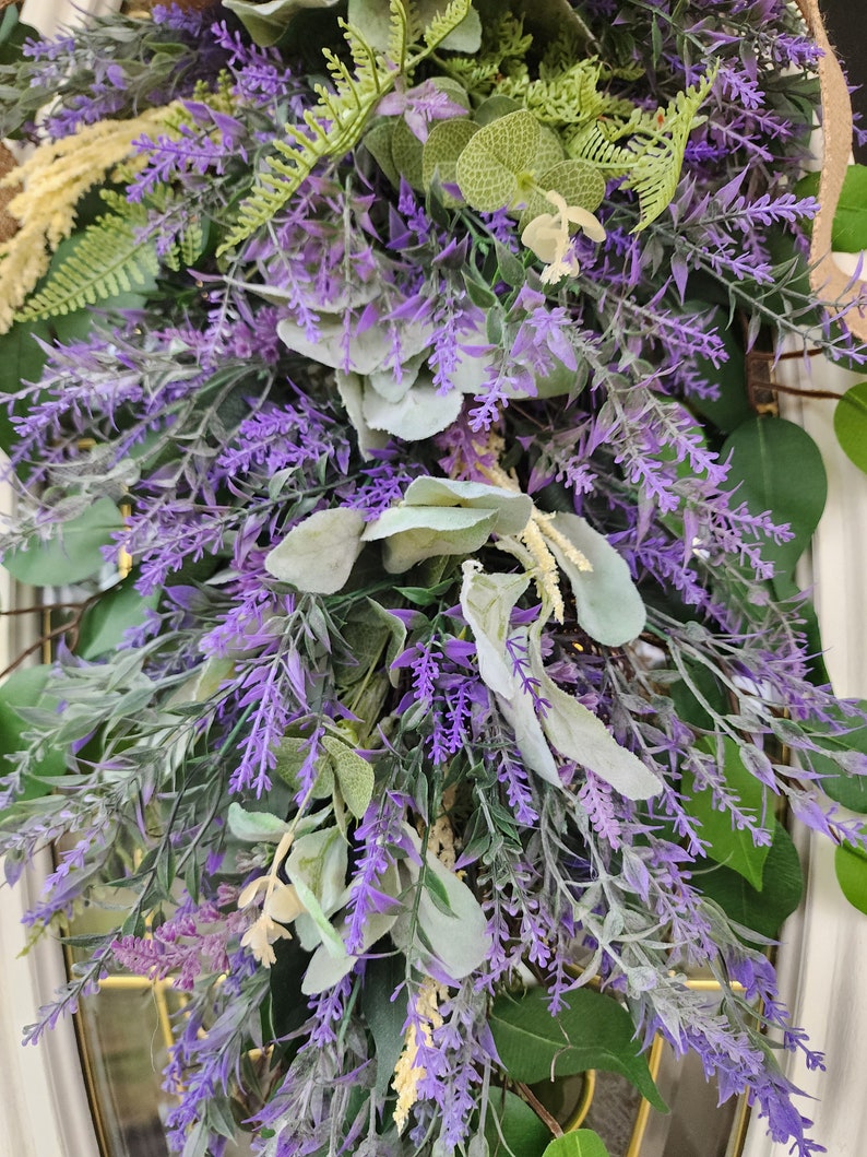 Peut inclure: Gros plan sur une couronne d&eacute;corative avec de la lavande violette, des feuilles vertes et un feuillage cr&egrave;me. La couronne est dens&eacute;ment garnie de diverses textures et nuances de vert et de violet, cr&eacute;ant une esth&eacute;tique naturelle et accueillante.