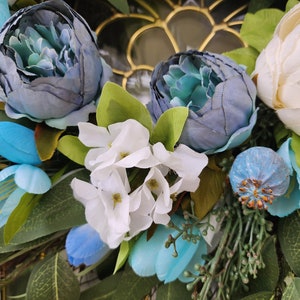 May include: A wreath made of artificial flowers in shades of blue and white. The wreath features large blue peonies, smaller blue flowers, and white hydrangeas. The wreath is accented with green leaves.