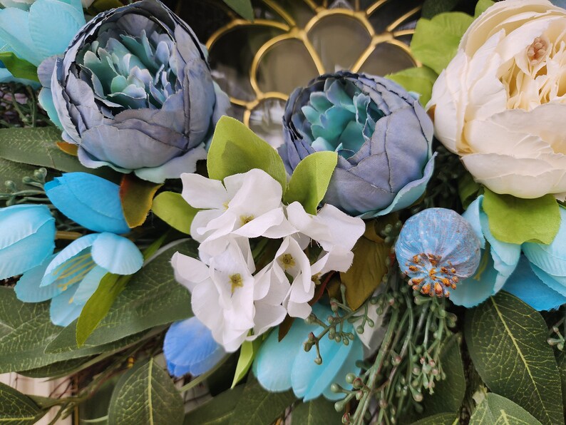 May include: A wreath made of artificial flowers in shades of blue and white. The wreath features large blue peonies, smaller blue flowers, and white hydrangeas. The wreath is accented with green leaves.